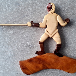 Puede incluir: Una figura de madera de un esgrimista en una pose de esgrima, sosteniendo una espada. La figura está hecha de madera clara y oscura y está de pie sobre una base de madera.