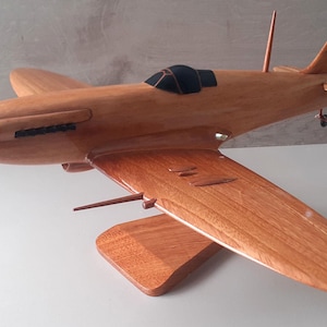 Puede incluir: Un modelo de avión de madera con hélice, alas y cola. El avión es de madera marrón claro y tiene una cabina negra. El modelo se exhibe sobre un soporte de madera.