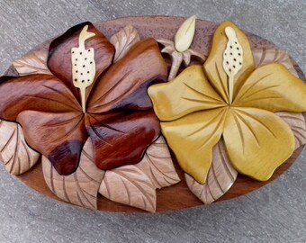 May include: A wooden puzzle box featuring a carved design of two hibiscus flowers. The flowers are in shades of brown and yellow, and the leaves are a light brown.