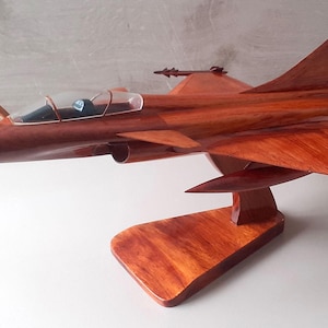 Pode incluir: Modelo em madeira de um avião de combate a jato, com asas, barbatanas de cauda e cockpit. O modelo é montado em um suporte de madeira e o avião é finalizado com um tom marrom avermelhado. Mede cerca de 40 cm de comprimento.