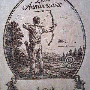 Può includere: Una targa in legno chiaro con un'illustrazione incisa al laser scuro di un arciere che mira a un bersaglio in un campo. Il testo recita Bon Anniversaire e Âge, con un nastro vuoto in basso per la personalizzazione e una piccola icona a bersaglio.