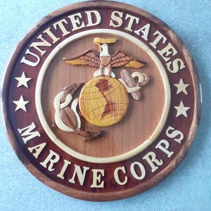Peut inclure: Une plaque en bois avec l'emblème du Corps des Marines des États-Unis. L'emblème représente un globe surmonté d'un aigle, entouré d'une corde et d'une ancre. Les mots "United States Marine Corps" sont inscrits autour de l'emblème.