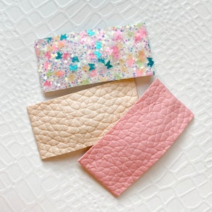 May include: Three rectangular hair clips. One is white with glitter and colorful butterfly shapes. The other two are textured, one in a light beige and the other in a soft pink.