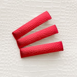 May include: Three bright red rectangular hair clips with a textured surface are arranged diagonally on a white, patterned background. The clips are made of a fabric-like material and appear to be new.