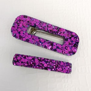 May include: Two hair clips featuring a vibrant purple and dark blue chunky glitter finish. The larger clip is a rectangular frame style with a silver metal snap mechanism, while the smaller clip is a solid rectangular bar with a silver metal base.