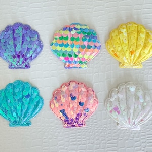 May include: Six colorful seashell-shaped patches with a sparkly, iridescent finish. The shells are in shades of purple, green, yellow, pink, and white.
