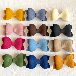 May include: Twelve felt hair bows in various colors, including pink, blue, brown, green, and yellow. The bows are arranged in three rows of four bows each.