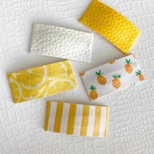 May include: Five different hair clips with a summer theme. The clips are rectangular and feature various patterns, including lemons, pineapples, stripes, and solid colors. The clips are all yellow and white.