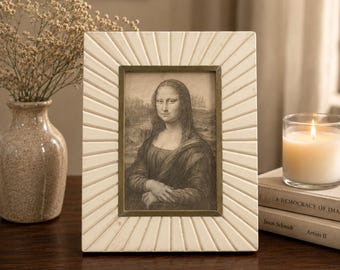 Puede incluir: Una impresión enmarcada de la Mona Lisa. El marco es de color blanco roto con un diseño de rayos de sol y un marco interior de color latón. La impresión es en blanco y negro. También se ven una vela encendida y un jarrón con flores secas.