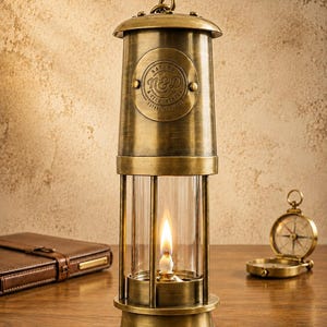 Op de afbeelding: Een koperen mijnwerkerslamp met een cilindrisch glazen middenstuk, een brandende lont en een ronde metalen plaat met de tekst Nature & Découvertes N&D Established 1995. De lamp heeft een handvat aan de bovenzijde en een stevige voet.
