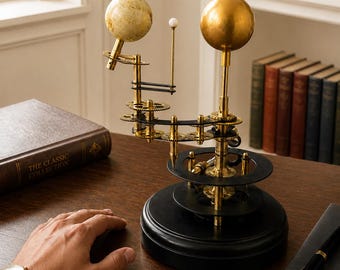 Op de afbeelding: Een mechanisch orrery-model met een getextureerde gebroken witte wereldbol en een gladde goudkleurige bol, gemonteerd op een messing armconstructie met zichtbare tandwielen. Het apparaat rust op een ronde zwarte voet en bevat een kleine witte verticale staaf.