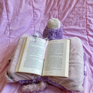 May include: A purple and white plush pillow with a book resting on top. The book is open to a page with the title "Firestorm" and the text "The Goblin Prince said."