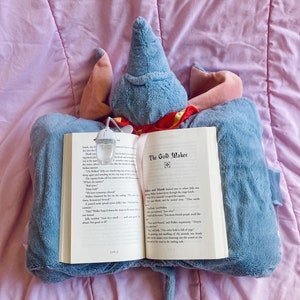 May include: A blue and red plush toy is holding an open book titled "The Gold Maker". The book is open to a page with text about a character named "Jolly" and a description of the character's actions.