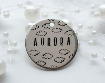 May include: A silver round tag with a cloud pattern and the name "AURORA" engraved in black lettering.