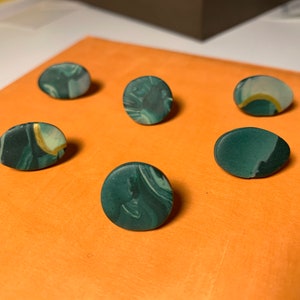 May include: Six handcrafted polymer clay earrings in shades of green and white, with gold detailing. The earrings are in various shapes, including round and oval, and are displayed on an orange surface.