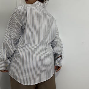 May include: A long-sleeved button-down shirt with vertical stripes in light blue, white, and tan. The shirt has a classic collar and buttoned cuffs. The fabric appears to be a lightweight cotton blend.