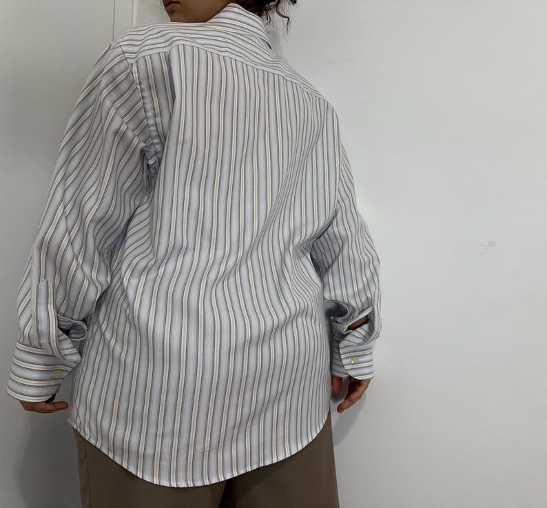 May include: A long-sleeved button-down shirt with vertical stripes in light blue, white, and tan. The shirt has a classic collar and buttoned cuffs. The fabric appears to be a lightweight cotton blend.