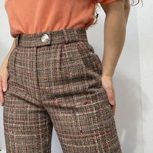 May include: High-waisted brown tweed pants with a subtle plaid pattern in red, white, and orange. The pants feature a button closure and pleats at the waist. The fabric appears to be a woven tweed, suitable for cooler weather.