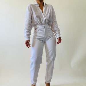 May include: A light-colored linen button-down shirt and white high-waisted jeans. The jeans have visible stitching and a red tag on the back pocket. The outfit is styled with a relaxed fit.