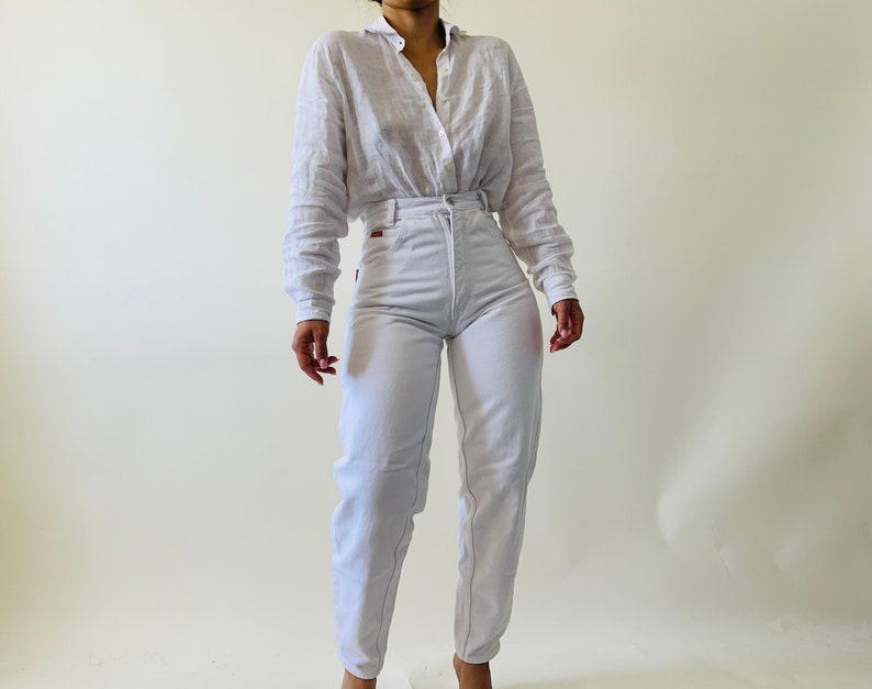 May include: A light-colored linen button-down shirt and white high-waisted jeans. The jeans have visible stitching and a red tag on the back pocket. The outfit is styled with a relaxed fit.