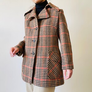 May include: A vintage coat with a classic houndstooth pattern in tan, black, and red. The coat features a notched collar, button closure, and two front pockets. The coat is a mid-length style.