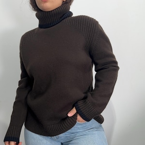 May include: A brown turtleneck jumper with ribbed detailing on the shoulders and cuffs. The collar and cuffs have a contrasting black trim. The jumper is made of a soft, knitted material.