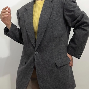 Könnte beinhalten: Ein anthrazitfarbener Blazer mit klassischem Zwei-Knopf-Verschluss und fallendem Revers. Der Blazer hat zwei Vordertaschen und geknöpfte Manschetten. Ein gelber Pullover ist darunter sichtbar.