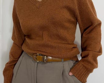 May include: A rust-colored V-neck sweater with long sleeves. The sweater is paired with gray trousers and a brown leather belt with a gold buckle. The person is wearing the sweater and trousers.