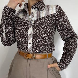 May include: A brown floral print shirt with long sleeves and a collared neckline. The shirt features a contrasting plaid and solid panel design on the yoke and cuffs. A brown leather belt with a gold buckle is worn with the shirt.