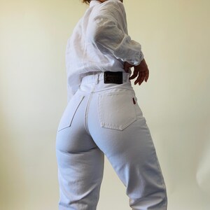 May include: White high-waisted jeans with a black leather label that reads "Annabella's" and a red tag on the back pocket. The jeans are paired with a white long-sleeved shirt.