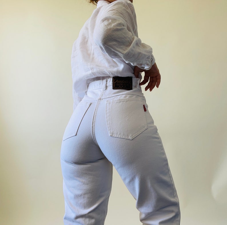 May include: White high-waisted jeans with a black leather label that reads "Annabella's" and a red tag on the back pocket. The jeans are paired with a white long-sleeved shirt.