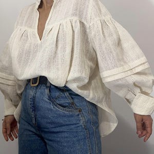 Puede incluir: Una blusa de manga larga en color blanco roto con un patr&oacute;n de rayas verticales texturizadas, cuello con volantes y escote en V profundo. La blusa presenta mangas voluminosas con pu&ntilde;os anchos y est&aacute; metida dentro de unos vaqueros azules con cintur&oacute;n.