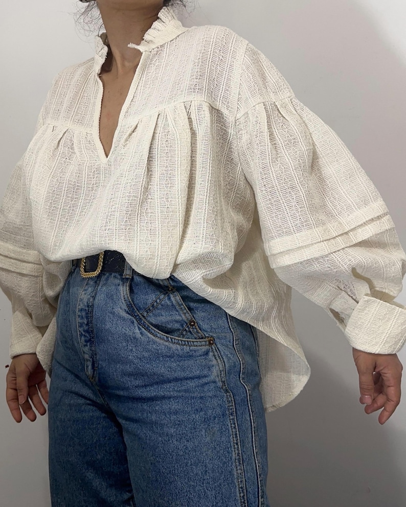 Puede incluir: Una blusa de manga larga en color blanco roto con un patr&oacute;n de rayas verticales texturizadas, cuello con volantes y escote en V profundo. La blusa presenta mangas voluminosas con pu&ntilde;os anchos y est&aacute; metida dentro de unos vaqueros azules con cintur&oacute;n.
