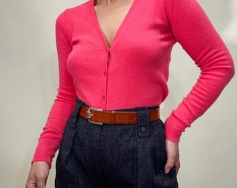 May include: A bright pink cardigan with a V-neck and button closure. The cardigan is paired with dark gray high-waisted trousers and a brown leather belt with a silver buckle. The outfit is a classic and stylish ensemble.