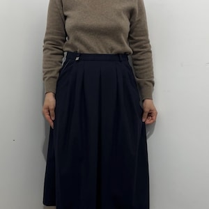 May include: A beige long-sleeve turtleneck sweater paired with a dark navy blue midi-length skirt. The skirt features a structured waistband with a small silver-toned button detail and vertical pleats that create a full, flowing silhouette.
