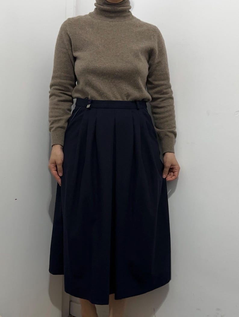 May include: A beige long-sleeve turtleneck sweater paired with a dark navy blue midi-length skirt. The skirt features a structured waistband with a small silver-toned button detail and vertical pleats that create a full, flowing silhouette.