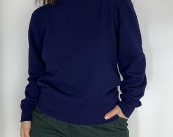 May include: A navy blue sweater with a crew neck and long sleeves. The sweater is made of a soft material and is a solid color. The person is wearing green pants.