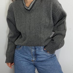 May include: A charcoal gray long-sleeve sweater featuring a V-neckline with a white and black striped trim detail. The sweater has a ribbed texture throughout the body and sleeves, paired with blue denim jeans that have a button-fly closure.