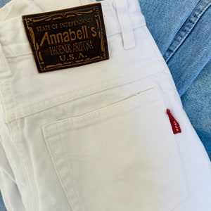May include: Close-up of white denim jeans with a brown leather label that reads "STATE OF INDEPENDENCE Annabell's PHOENIX ARIZONA U.S.A." A small red tag is attached to the back pocket. The jeans are folded and rest on top of a light blue denim.
