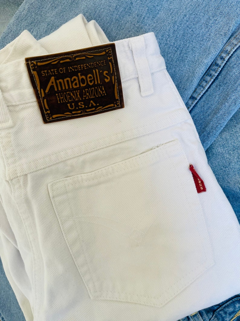 May include: Close-up of white denim jeans with a brown leather label that reads "STATE OF INDEPENDENCE Annabell's PHOENIX ARIZONA U.S.A." A small red tag is attached to the back pocket. The jeans are folded and rest on top of a light blue denim.