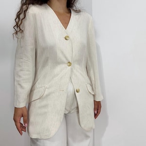 May include: A cream-colored blazer with a V-neck and three round, light-colored buttons. The blazer has two front pockets and long sleeves. The fabric has a subtle textured pattern. The blazer is paired with white pants.