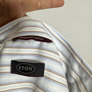 May include: Close-up of a light blue and white striped shirt with brown and blue accent stripes. The shirt has a label that reads "ETON" and a tag that says "wrinklefree cotton".