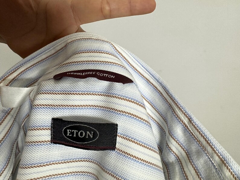 May include: Close-up of a light blue and white striped shirt with brown and blue accent stripes. The shirt has a label that reads "ETON" and a tag that says "wrinklefree cotton".