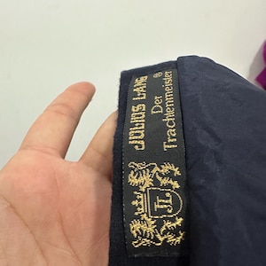 May include: A dark navy blue garment featuring a black interior label with gold embroidery. The label displays the text Julius Lang Der Trachtenmeister and a crest featuring a shield with the letters JL flanked by two lions and a crown.
