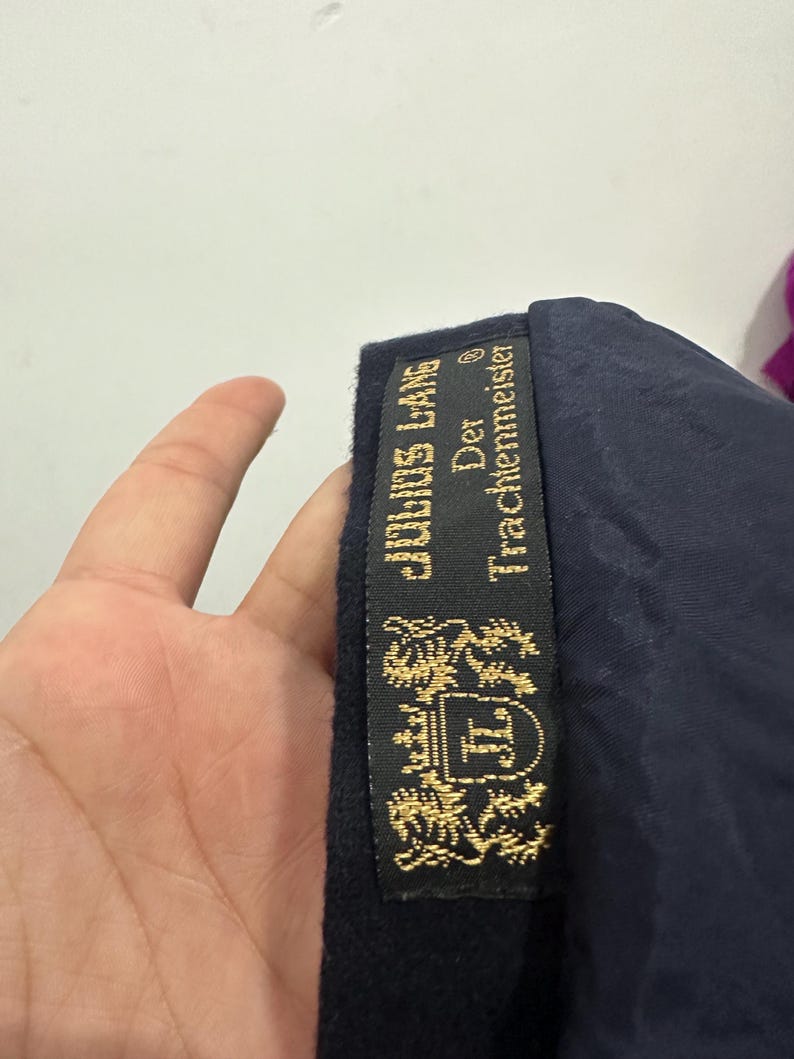 May include: A dark navy blue garment featuring a black interior label with gold embroidery. The label displays the text Julius Lang Der Trachtenmeister and a crest featuring a shield with the letters JL flanked by two lions and a crown.
