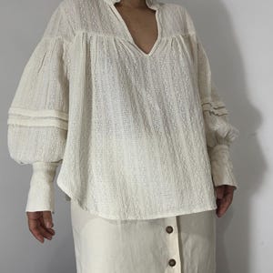 Puede incluir: Una blusa de manga larga en color blanco roto con un patr&oacute;n de rayas verticales texturizadas, cuello alto con volantes, escote en V y mangas voluminosas con pu&ntilde;os ajustados. Combinada con una falda en color blanco roto con botones marrones.