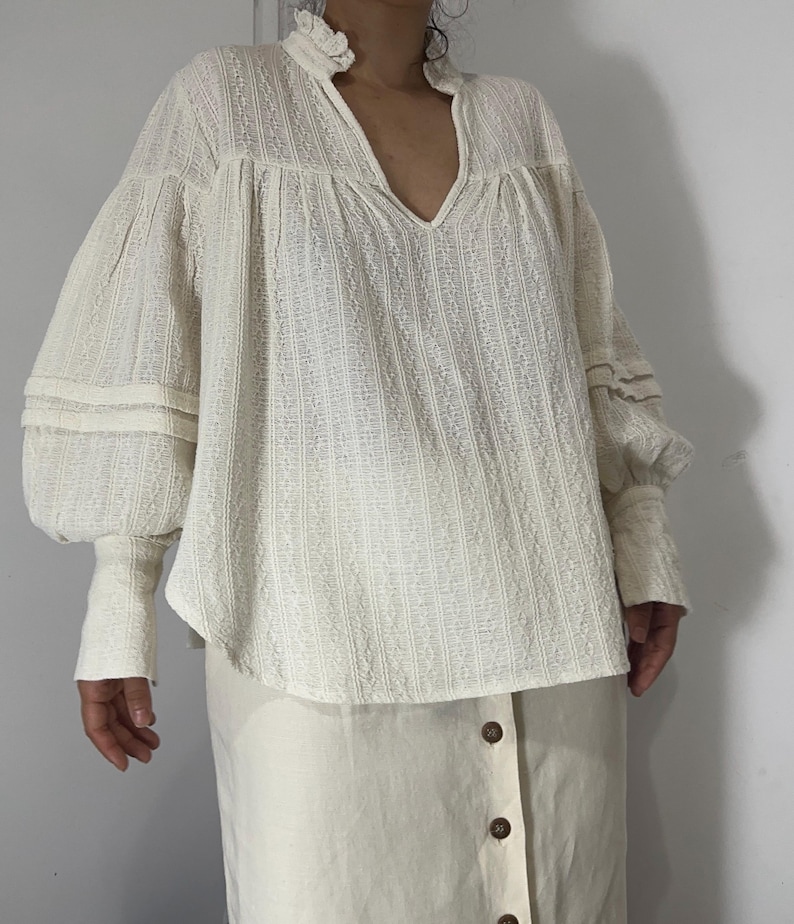 Puede incluir: Una blusa de manga larga en color blanco roto con un patr&oacute;n de rayas verticales texturizadas, cuello alto con volantes, escote en V y mangas voluminosas con pu&ntilde;os ajustados. Combinada con una falda en color blanco roto con botones marrones.