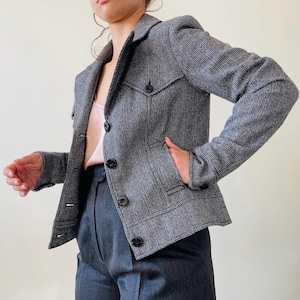 Puede incluir: Una blazer de tweed gris con solapa de muesca, dos bolsillos delanteros y botones negros. La blazer se combina con pantalones grises. La chaqueta tiene un diseño clásico y a medida.