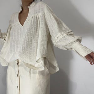 Puede incluir: Una blusa de manga larga color blanco hueso con tejido texturizado de rayas verticales, escote en V con cuello de volantes y mangas voluminosas con pu&ntilde;os ajustados, combinada con una falda color blanco hueso con botones marrones.