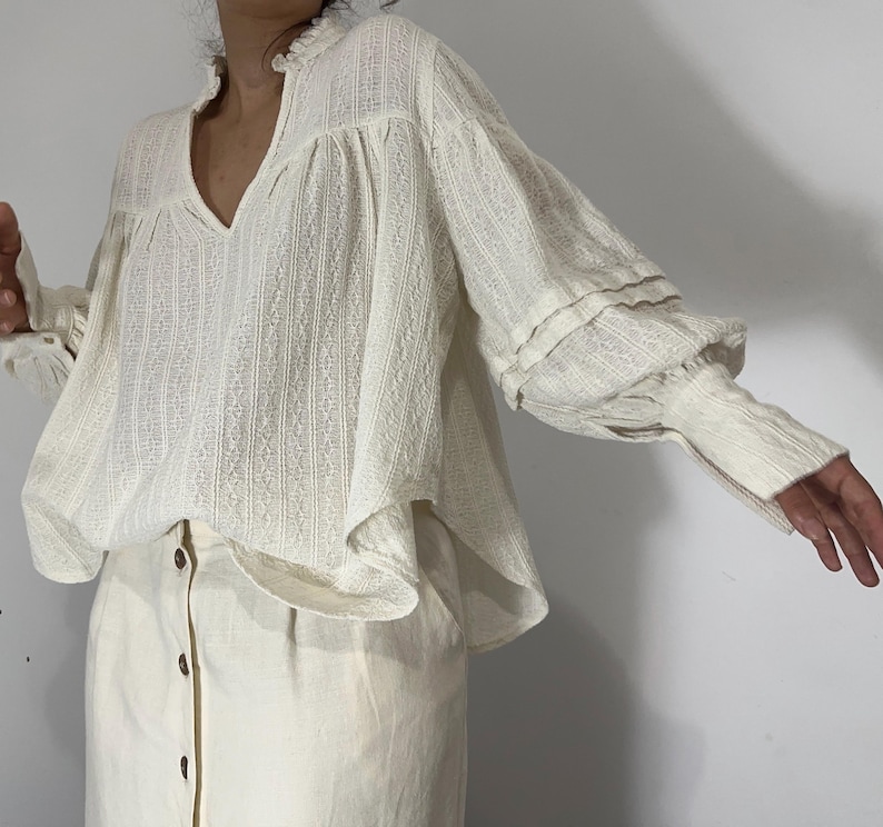 Puede incluir: Una blusa de manga larga color blanco hueso con tejido texturizado de rayas verticales, escote en V con cuello de volantes y mangas voluminosas con pu&ntilde;os ajustados, combinada con una falda color blanco hueso con botones marrones.